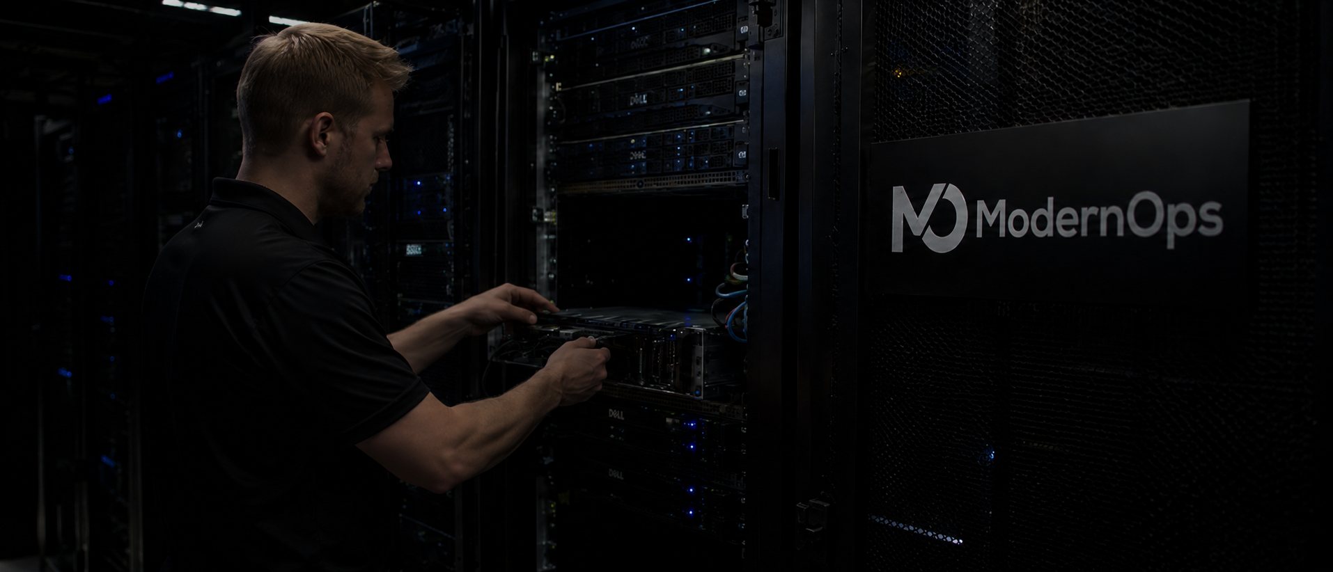 ModernOps engineer cabling a Dell PowerEdge node in a rack on the ModernOps data center floor, ModernOps logo on the cage door